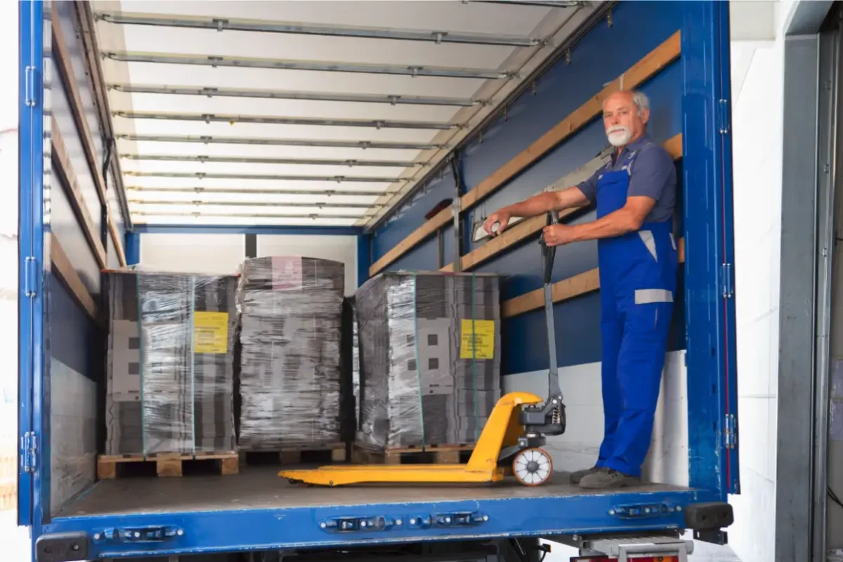An older man in blue overalls uses a pallet jack to move shrink-wrapped pallets inside the back of a blue delivery truck. Traffic Consultants, Inc. (TCI)