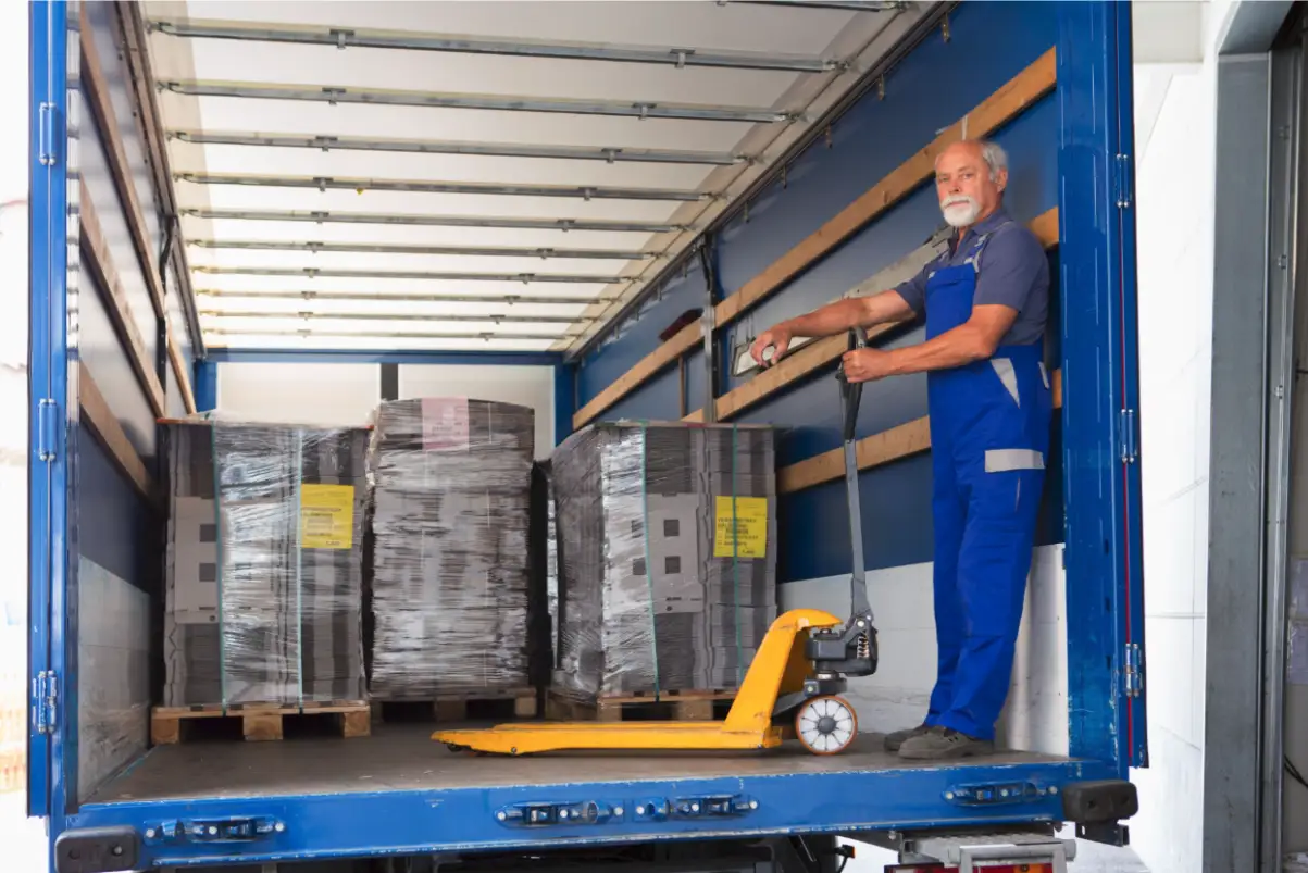 An older man in blue overalls uses a pallet jack to move shrink-wrapped pallets inside the back of a blue delivery truck. Traffic Consultants, Inc. (TCI)