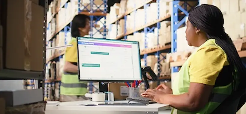 A woman in a yellow high-visibility vest works at a computer in a warehouse, while another person walks among shelves filled with boxes. Traffic Consultants, Inc. (TCI)