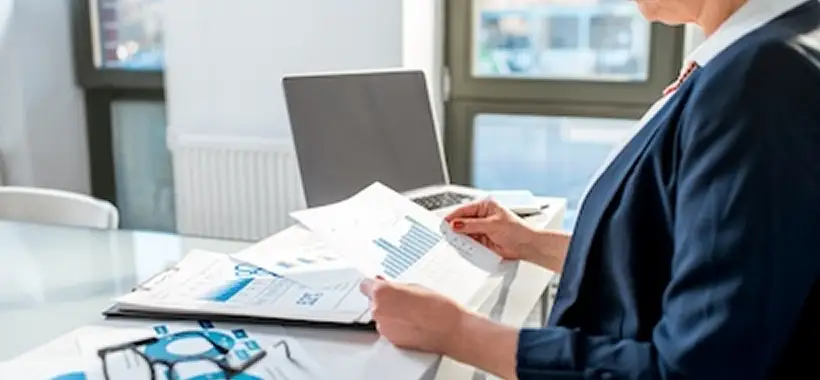 Person in business attire reviewing graphs and charts at a desk with a laptop, papers, and eyeglasses. Bright office setting with large windows in the background. Traffic Consultants, Inc. (TCI)