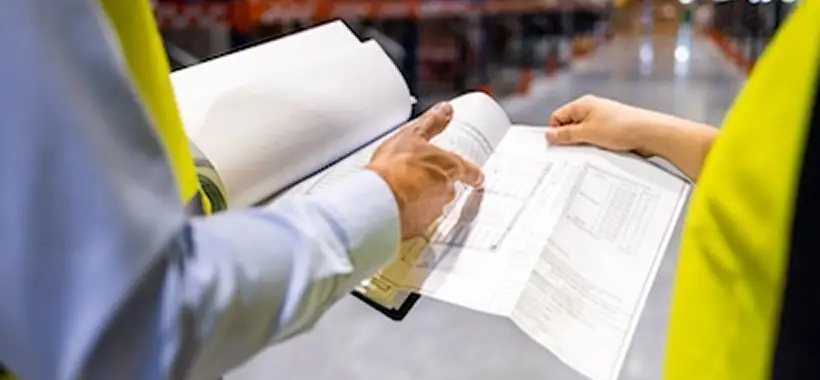 Two people in safety vests review documents on clipboards in a warehouse setting, focusing on paperwork and inventory lists. Traffic Consultants, Inc. (TCI)