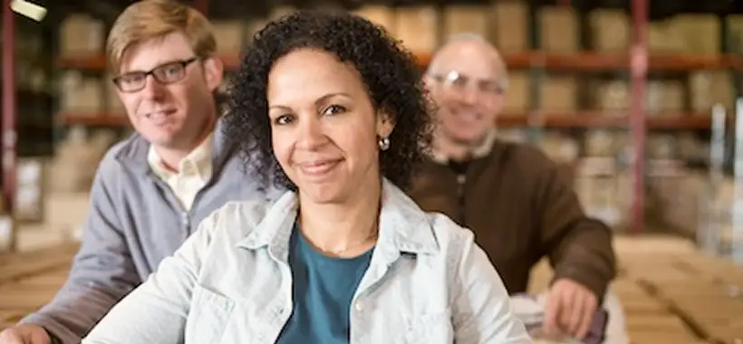 Three people pose together in a warehouse, with shelves of boxes in the background; the woman in front smiles at the camera. Traffic Consultants, Inc. (TCI)