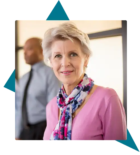 Older woman with short gray hair, wearing a pink sweater and patterned scarf, stands indoors with a blurred man in business attire in the background. Traffic Consultants, Inc. (TCI)