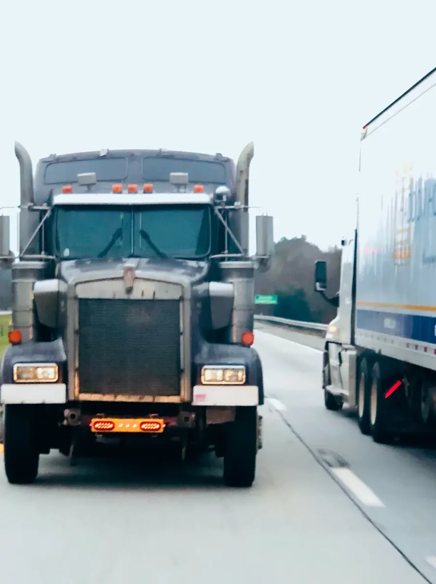 A dark semi-truck drives on a highway next to a white trailer truck under a cloudy sky. Traffic Consultants, Inc. (TCI)