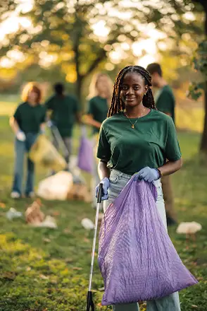 A woman in a green shirt holds a purple trash bag and a litter picker, cleaning up a park with a group of people in the background. Traffic Consultants, Inc. (TCI)