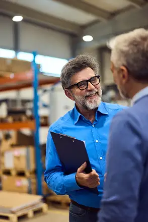 A man in a blue shirt holding a clipboard talks to another man in a warehouse setting with shelves and boxes in the background. Traffic Consultants, Inc. (TCI)