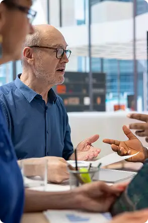 Three people sit at a table in an office, engaged in discussion. One older man gestures with his hands while others listen and hold documents and a tablet. Traffic Consultants, Inc. (TCI)