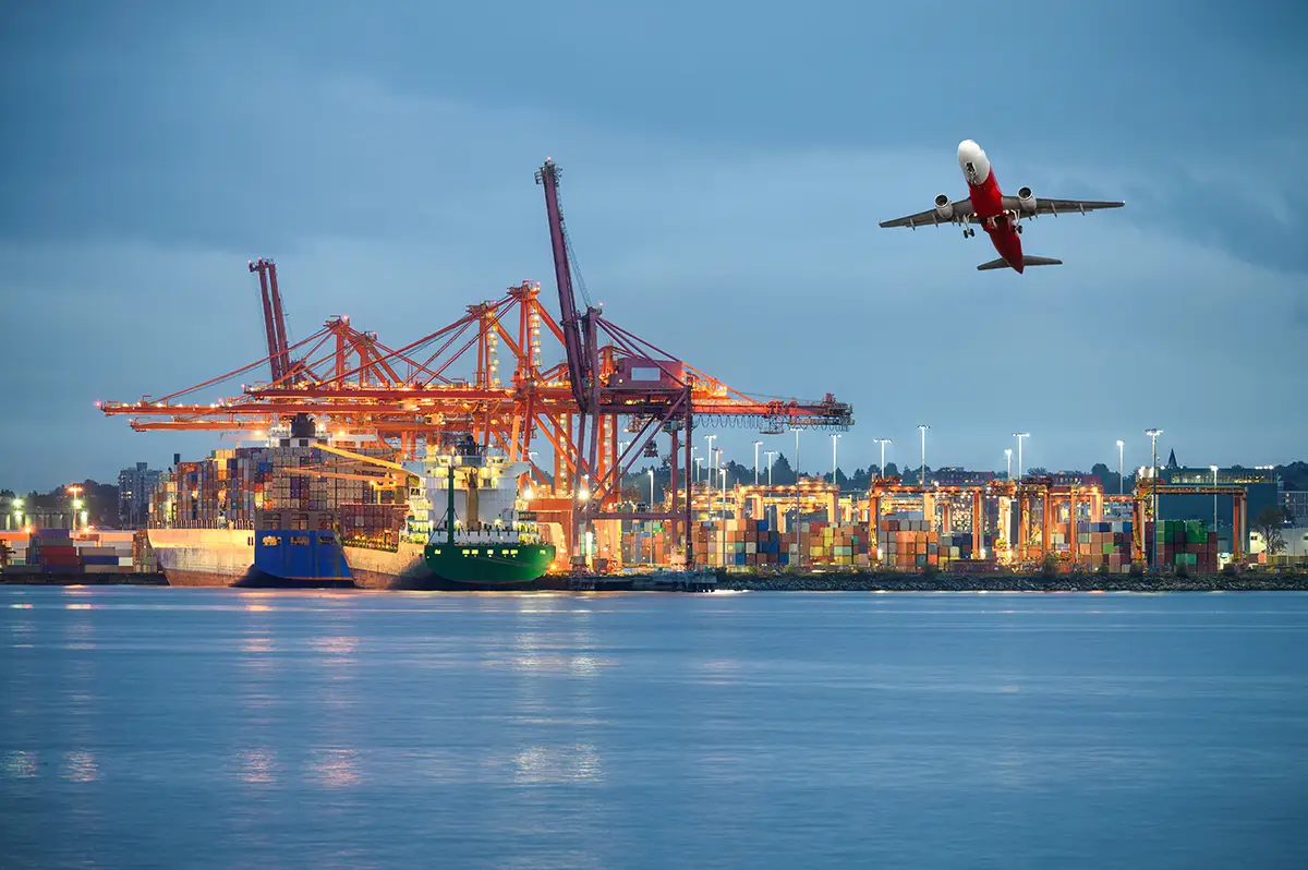 A cargo ship docked at a busy port with cranes, colorful shipping containers, and an airplane taking off overhead at dusk. Traffic Consultants, Inc. (TCI)